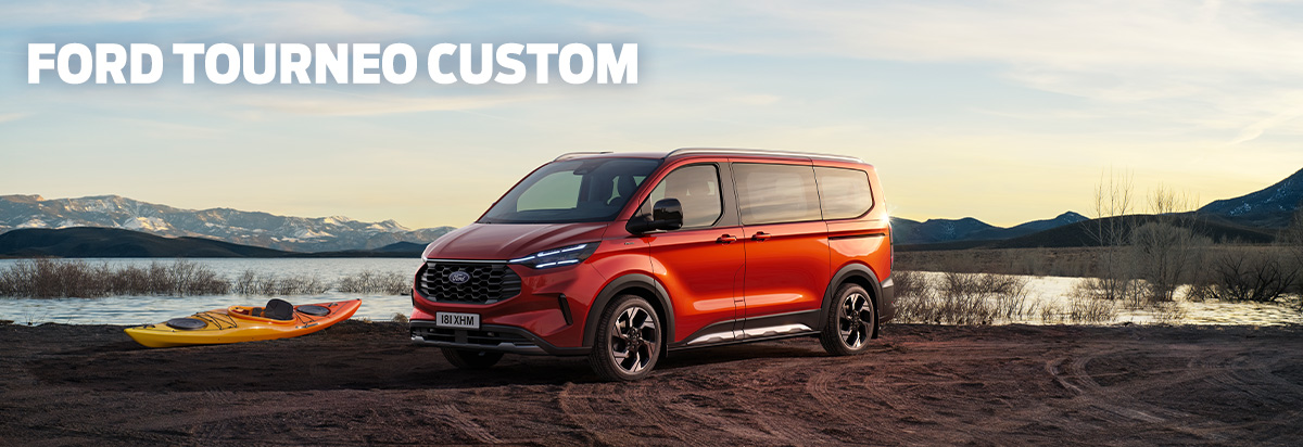 Ford Tourneo Custom in Orange, parkt am Ufer eines Gewässers mit Bergen im Hintergrund, Kajak liegt am Strand.